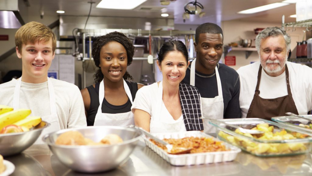 Diverse group of volunteers in kitchen smiling with meal they all helped to prepare